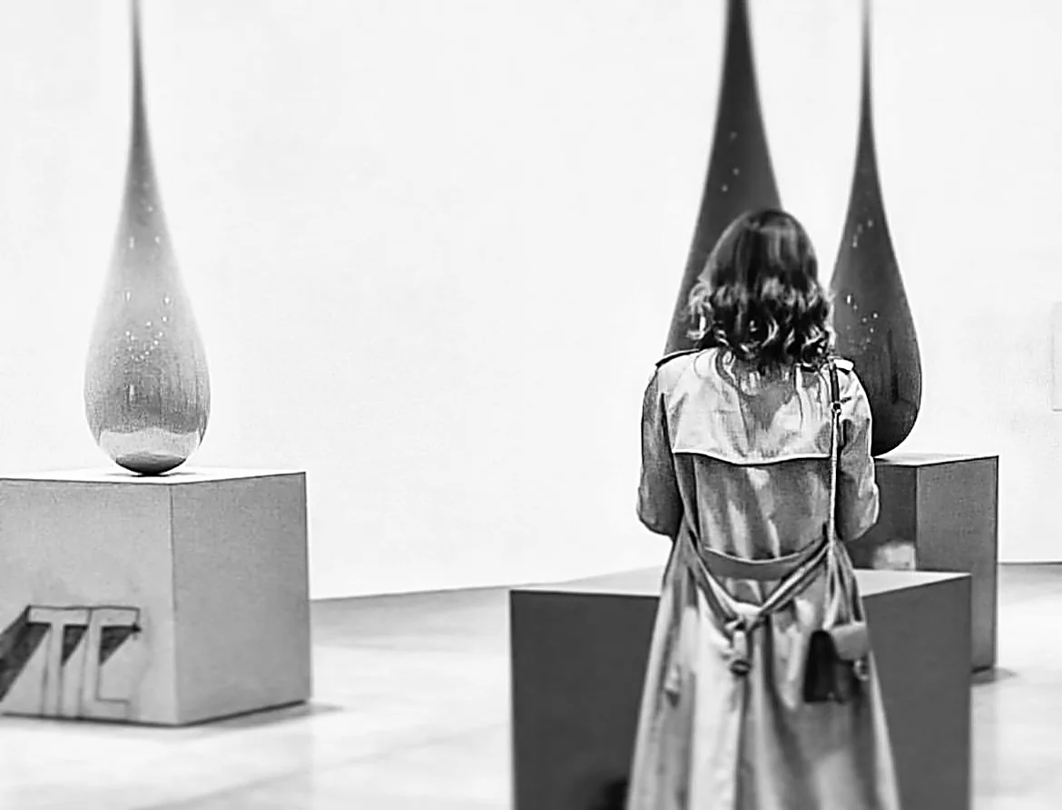 Black and white photograph of Gabriella Vittoria Monti seen from behind, contemplating a contemporary sculpture in a gallery