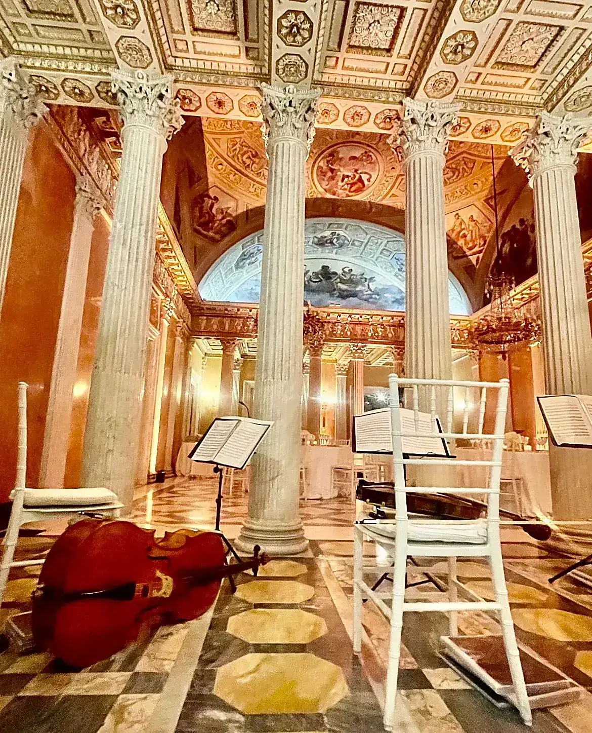 Frescoed hall of a historic Italian palazzo prepared for a private concert, with a cello resting between marble columns